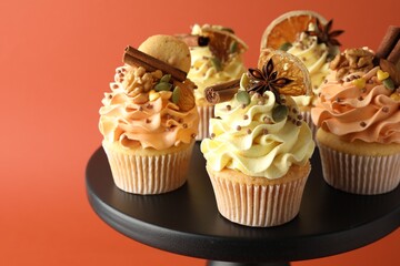 Delicious cupcakes with sprinkles, cookies, spices and dry orange slices on stand against coral background, closeup. Autumn atmosphere