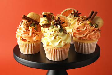 Delicious cupcakes with sprinkles, cookies, spices and dry orange slices on stand against coral background, closeup. Autumn atmosphere