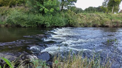 Cinematic low angle view of the River Maine at Galgorm, Northern Ireland, with flowing water over rocks and lush vegetation - ideal for film, documentary, and nature productions.
