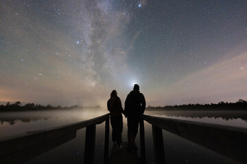 A romantic couple on a wooden pier enjoys the magical starry night sky with the Milky Way in a tranquil Estonian bog.