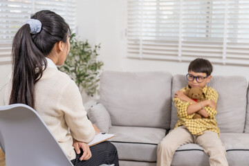 Young child sitting on sofa hugging teddy bear tightly, avoiding eye contact with therapist during counseling session, showing closed posture and emotional withdrawal in private consultation room