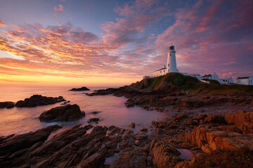 Lighthouse on rocky cliff at dramatic sunset by the ocean.