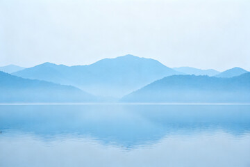 Calm lake reflecting misty mountains under a soft blue sky