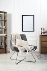 Stylish armchair with pillow, blanket, dresser and shelving unit indoors. Interior design