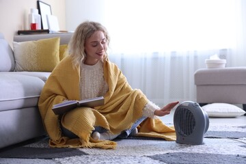 Young woman reading book near electric heater at home