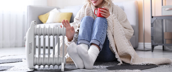 Young woman with cup of drink near electric heater on floor at home, closeup