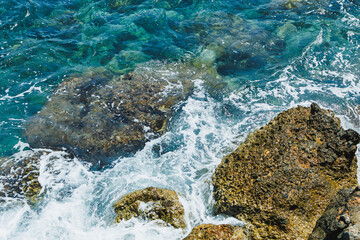 Waves crash against rugged stones in a vibrant turquoise sea under a bright sunny sky