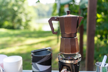 Brown moka pot coffee maker brewing on portable gas burner with white cup and camping equipment on outdoor table. Concept of camping coffee, outdoor breakfast and wilderness morning ritual preparation