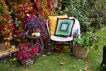 Cozy autumn chair with colorful pillows, crochet cushion, and seasonal plants in baskets. Surrounded by vibrant foliage and rustic garden decor, it creates a warm outdoor atmosphere perfect for relax