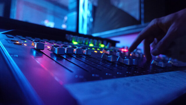 Close-up of a sound engineer's hand adjusting faders on a professional audio mixing console, lit by vibrant blue and purple neon lights. Capturing the dynamic atmosphere of a live event or studio sess