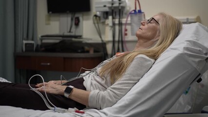 A mature woman is in a hospital room hooked up to heart and EKG monitors. She appears calm while receiving medical attention in the emergency department. - Powered by Adobe