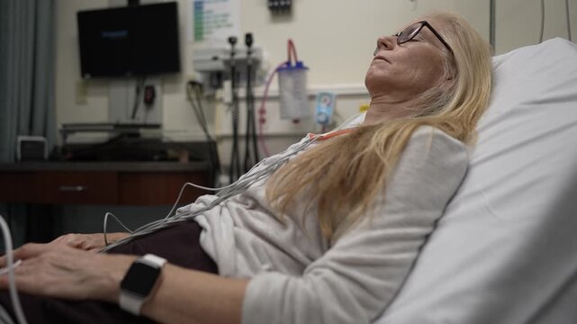 A mature woman is resting in an emergency hospital room while connected to heart monitors and EKG devices, receiving essential health care and monitoring.