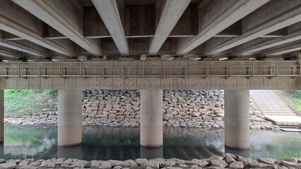 underneath a concrete bridge with piers over a stream and stone embankment