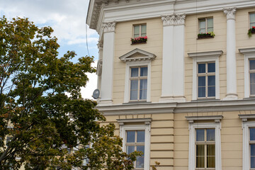 Elegant neoclassical building facade with ornate Corinthian columns, decorative pediments and flower boxes against green foliage. Concept of European architectural heritage and urban elegance.