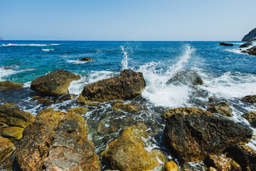 Serene waves crashing against rugged rocks at the sunny coastline of a secluded beach under a clear blue sky