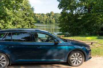 Dark blue modern SUV car parked by serene lake surrounded by lush green forest on sunny summer day. Concept of road trip, nature travel, outdoor adventure and automotive lifestyle.