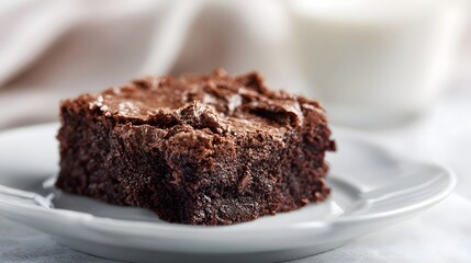 A rich dark chocolate brownie sits on a clean white plate with a blurred background of milk and fabric