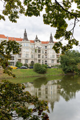 Obraz premium Elegant historic mansion with ornate towers and red tile roofs reflected in calm river water framed by green tree branches. European architectural heritage, luxury real estate and waterfront property.