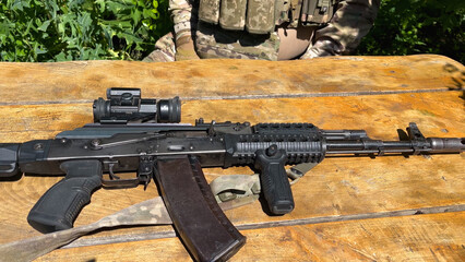 Kalashnikov assault rifle lies on the table. A soldier in uniform is sitting next to the weapon....