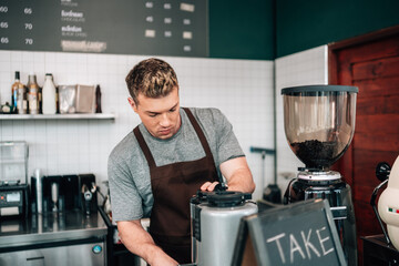 male barista in a brown apron is working behind the counter, operating a piece of coffee equipment....