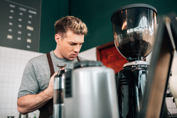 male barista in a brown apron is working behind the counter, operating a piece of coffee equipment. A grinder filled with coffee beans is visible to his right.