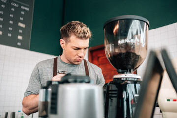 male barista in a brown apron is working behind the counter, operating a piece of coffee equipment. A grinder filled with coffee beans is visible to his right.
