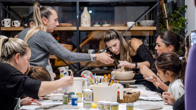 In an art studio during a group art event, a group of women and children are enjoying a creative hobby, learning and having fun as they make shapes out of clay - Powered by Adobe