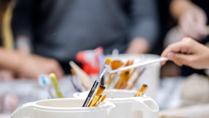 A close-up shot of paintbrushes in the foreground, with two women artists working on handmade products in the background of an art studio