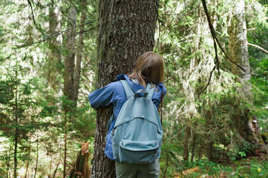 Young blonde woman with a pastel blue backpack hugging a tree in a lush green forest. Peaceful moment of connection with nature, surrounded by tall trees and soft forest light