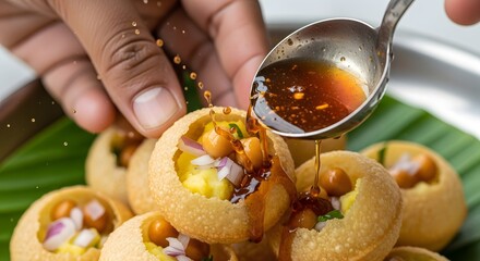 A hand drizzles tangy sauce over a plate of colorful, filled puris on a banana leaf backdrop