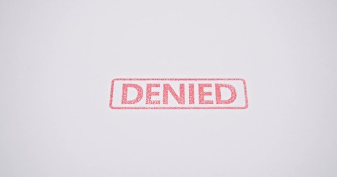 A video footage showing a close-up of a hand using a red rubber stamp to mark the word "DENIED" on a white sheet of paper. Representing refusal, rejection, or disapproval of applications or documents.