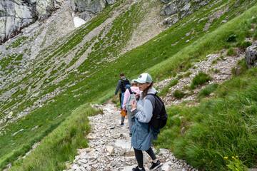  Woman with backpack hiking on stone mountain trail through lush green slopes in Tatra peaks with other hikers. Concept of solo female hiking, mountain trekking and independent outdoor adventure.