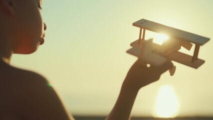 Happy childhood, little boy holding wooden aircraft model in hands and playing. Back view against bright sun in sky, sunset or sunrise above sea or ocean, cinematic slow motion, children game outdoors - Powered by Adobe