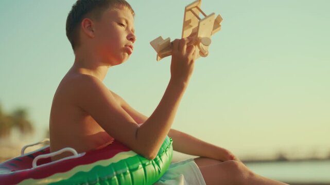 Child boy sitting alone on sandy beach on sea coast and playing with toy plane. Cinematic slow motion shot, bored kid viewing wooden aircraft model and dreaming of flight and traveling to new location