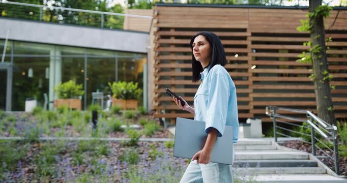 Side view of young Caucasian business woman in casual outfit use smartphone and holding laptop in hand while walking at front of modern building at city park. Business and people concepts