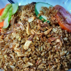 Close up of Indonesian fried rice is made with jasmine rice, shredded chicken, meatballs, eggs, and various green vegetables. It's topped with sliced tomatoes, cucumbers, and carrots. 