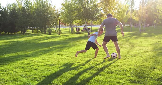 father and son play soccer on green grass of city park. sunlit trees and fresh atmosphere family interaction, energy time together outdoors. Father and son run around the sunlit grass ball.