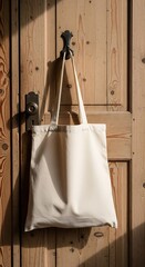 A blank off-white canvas tote bag hangs from a hook on a rustic wooden door, illuminated by warm sunlight.
