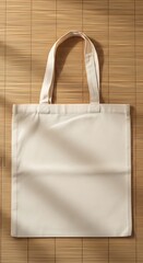 A plain, off-white canvas tote bag with two handles rests on a textured bamboo surface, casting soft shadows.
