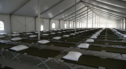Fototapeta premium Rows of cots with pillows fill a large, bright tent, possibly a shelter or temporary housing.