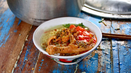 Chicken noodle soup served in a white bowl on a rustic wooden table, garnished with fresh vegetables and vibrant red sauce, creating a comforting and appetizing scene.