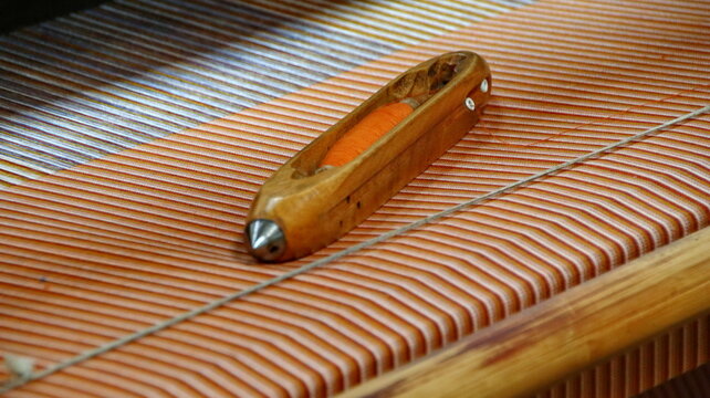 Wooden shuttle weaving orange and white threads on a loom in a textile factory, showcasing traditional craftsmanship and detailed texture. - Powered by Adobe