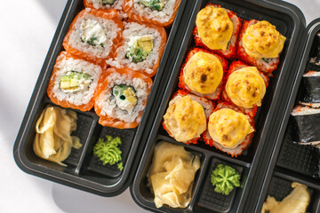Top view of assorted sushi rolls in black plastic container with wasabi and pickled ginger. Fresh salmon rolls with avocado and baked rolls with cheese topping. Japanese food delivery concept.