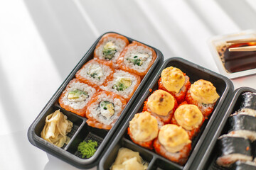 Assorted sushi set in plastic containers: salmon and avocado rolls, baked rolls with cheese topping. Overhead view with soy sauce and chopsticks in sunlight