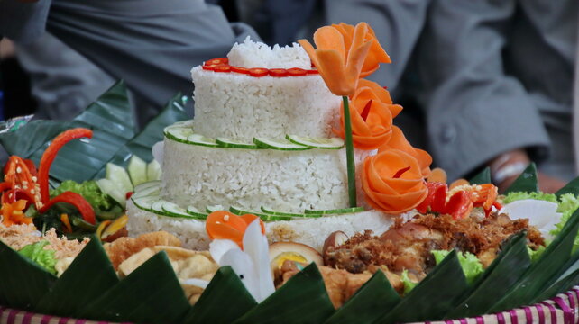 Tumpeng, a traditional Indonesian rice cone, sits decorated with vegetables and fried chicken on banana leaves during a celebration, showcasing cultural heritage.