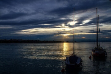 A picturesque view of sailboats moored on calm waters at sunset, with a dramatic cloudy sky and golden reflections. A peaceful seascape symbolizing travel, adventure, romance, and relaxation.