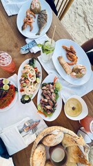 The image shows a table with various dishes, including fish, shrimp, salads, and bread. There are drinks and utensils, suggesting a restaurant setting. The background appears to be outdoors, possibly 