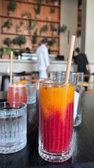 Two glasses of colorful drinks on a wooden table, surrounded by white sofas and palm trees, creating a relaxing outdoor atmosphere.