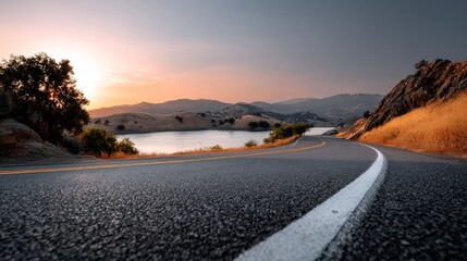 A winding road curves alongside a tranquil lake, framed by hills and bathed in warm sunset light, creating a serene landscape.