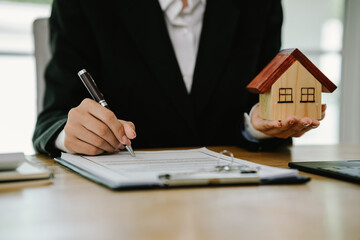 Businesswoman writing notes with house model, keys, contract, and calculator on desk, symbolizing property investment, real estate planning, and financial agreement.